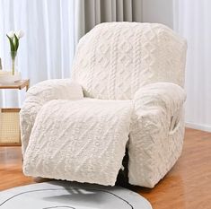 a white recliner covered in a blanket on top of a hard wood floor next to a vase with flowers