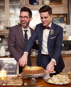 two men in suits cut into a cake on a table with other desserts around them