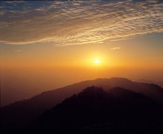 the sun is setting on top of a mountain with clouds in the sky above it