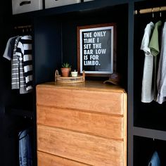 a wooden dresser sitting next to a black wall with clothes hanging on the walls and a framed poster above it