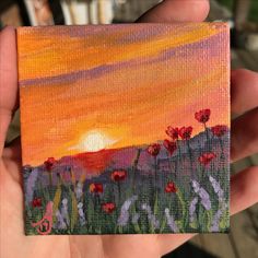 someone is holding up a small piece of art that has flowers painted on it and the sun setting in the background