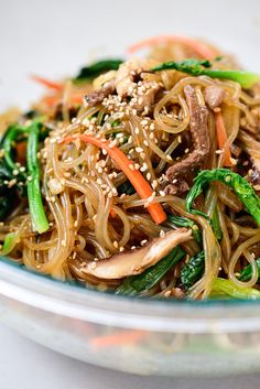 a glass bowl filled with noodles, meat and veggies covered in sesame seeds