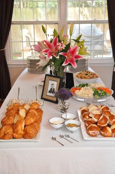 a table filled with lots of food on top of a white table cloth next to a window