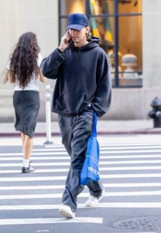 a man walking across a street talking on a cell phone while holding a blue bag