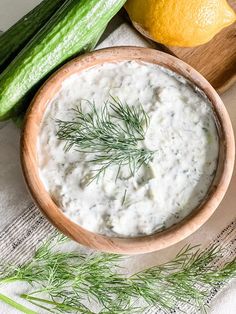 a wooden bowl filled with white sauce next to cucumbers and lemon wedges