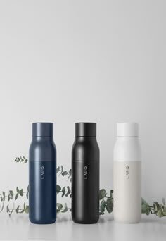 three different colored bottles sitting next to each other on a white surface with greenery