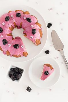 a pink cake with blackberries is on a plate next to a knife and fork