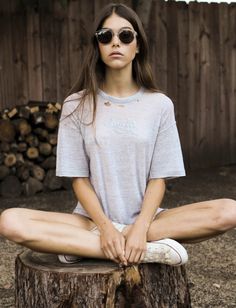 a woman sitting on top of a tree stump wearing sunglasses and a t - shirt