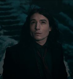 a man with long hair wearing a black suit and tie in front of a dark background