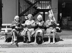 three old women sitting on a bench in front of a building, one holding a cup and the other looking at her cell phone