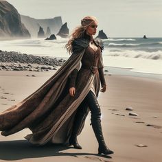 a woman is walking on the beach wearing a long dress and cape with her hands in her pockets