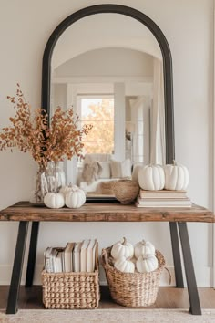 a wooden table topped with white pumpkins under a large mirror next to a basket filled with books