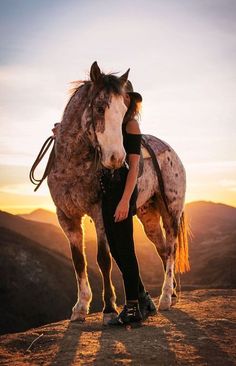 a woman standing next to a horse on top of a mountain at sunset or sunrise