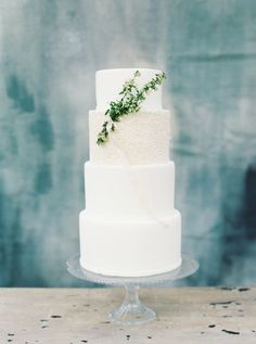 a three tiered white cake with green sprigs on top sits on a glass stand