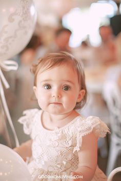 A baby girl with big, expressive eyes wearing a delicate lace dress sits amidst a festive setting, holding a white balloon while surrounded by blurred figures in the background, suggesting a celebration or party atmosphere.