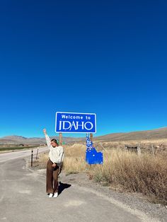 a man standing on the side of a road holding up a welcome to idaho sign