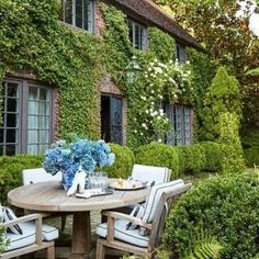 an outdoor dining table surrounded by greenery and blue hydrangeas in front of a house