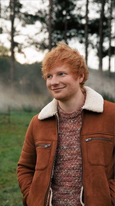 a man with red hair wearing a brown jacket and standing in front of some trees