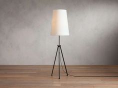 a floor lamp with a white shade on it in front of a gray wall and wooden floor