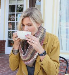 a woman sitting at a table drinking from a coffee cup while wearing a scarf around her neck