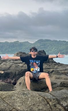 a man sitting on top of a large rock next to the ocean with his arms outstretched