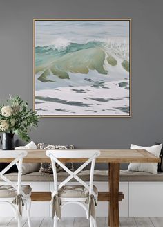 a painting hanging on the wall above a wooden table with white chairs and two vases filled with flowers