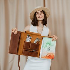 a woman in a hat is holding a book and some pens, pencils and scissors