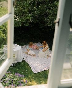 two women and a baby are sitting on a blanket in the grass near an open window