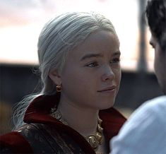 a woman with white hair wearing a red cape and gold earrings looking at another person