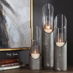 three candles are sitting in glass vases on a shelf next to books and a painting