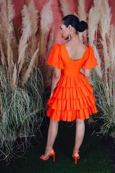 a woman in an orange dress standing next to some tall grass and dry grass with her back turned towards the camera