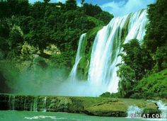 a large waterfall in the middle of a forest filled with green trees and water flowing down it's sides