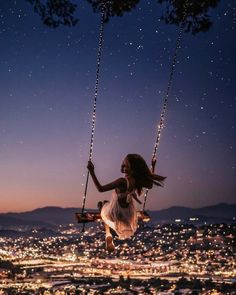 a woman is swinging on a swing in the air at night with city lights behind her