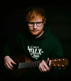 a man with glasses is playing an acoustic guitar in the dark while wearing a hoodie