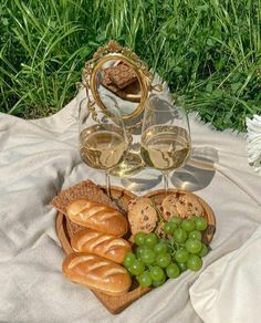 two glasses of wine, bread and grapes on a blanket with grass in the background