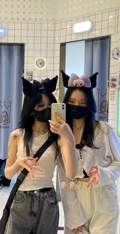 two young women wearing cat ears are looking at their cell phones while standing in front of a mirror