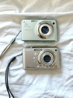 two cameras sitting on top of a white bed next to each other, one is silver and the other is black