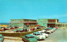 an old photo of cars parked in front of a building on the beach with people sitting at tables