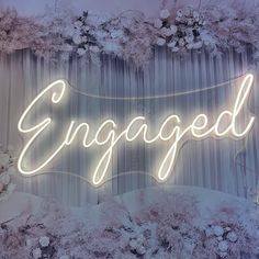 a neon sign that says engaged in front of curtains and flowers on the wall behind it