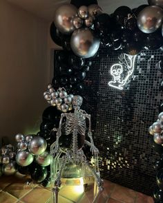 a skeleton sitting on top of a glass block in front of a wall with black and silver balloons