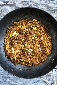 a wok filled with noodles and meat on top of a wooden table