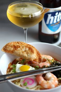 a white bowl filled with noodles and shrimp next to a glass of beer on top of a table