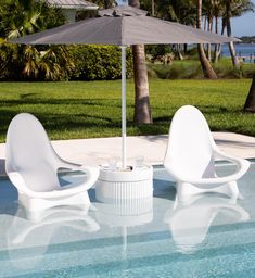 two white chairs sitting under an umbrella next to a swimming pool with grass and palm trees in the background