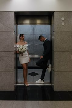 a man standing next to a woman in front of an open elevator with flowers on her head