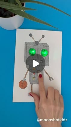 a person pointing at a paper cut out of a robot with green eyes and a coin