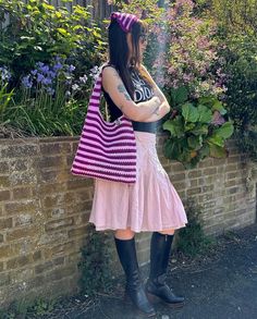 a woman in black boots and a pink dress holding a purple striped bag while standing next to a brick wall