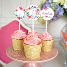 three cupcakes with pink frosting on a cake plate next to some flowers