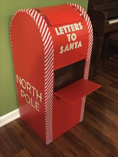 a red and white mailbox with letters to santa written on the front, next to a piano