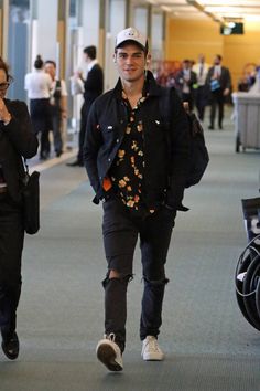 a man in black jacket and hat walking through an airport with other people behind him