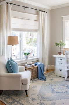 a white chair sitting in front of a window next to a table with a lamp on it